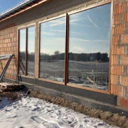 Montaż okien z brązowymi ramami w surowym budynku z cegły. Widoczne zimowe otoczenie i odbicie krajobrazu w szybach. Częściowo ośnieżony teren.