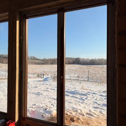 DMK - drzwi i okna z klimatem - Nowe okno z brązowymi ramami w surowym wnętrzu, widok na zimowy krajobraz z lasem w tle. Elementy montażowe widoczne u dołu ramy. Marka Gealan na profilach.