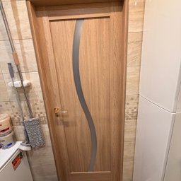 DMK - drzwi i okna z klimatem - Drewniane drzwi wewnętrzne z falistym, szarym przeszkleniem, widoczne w łazience z beżowymi płytkami. Złota klamka i fragment mopa dodają realizmu.