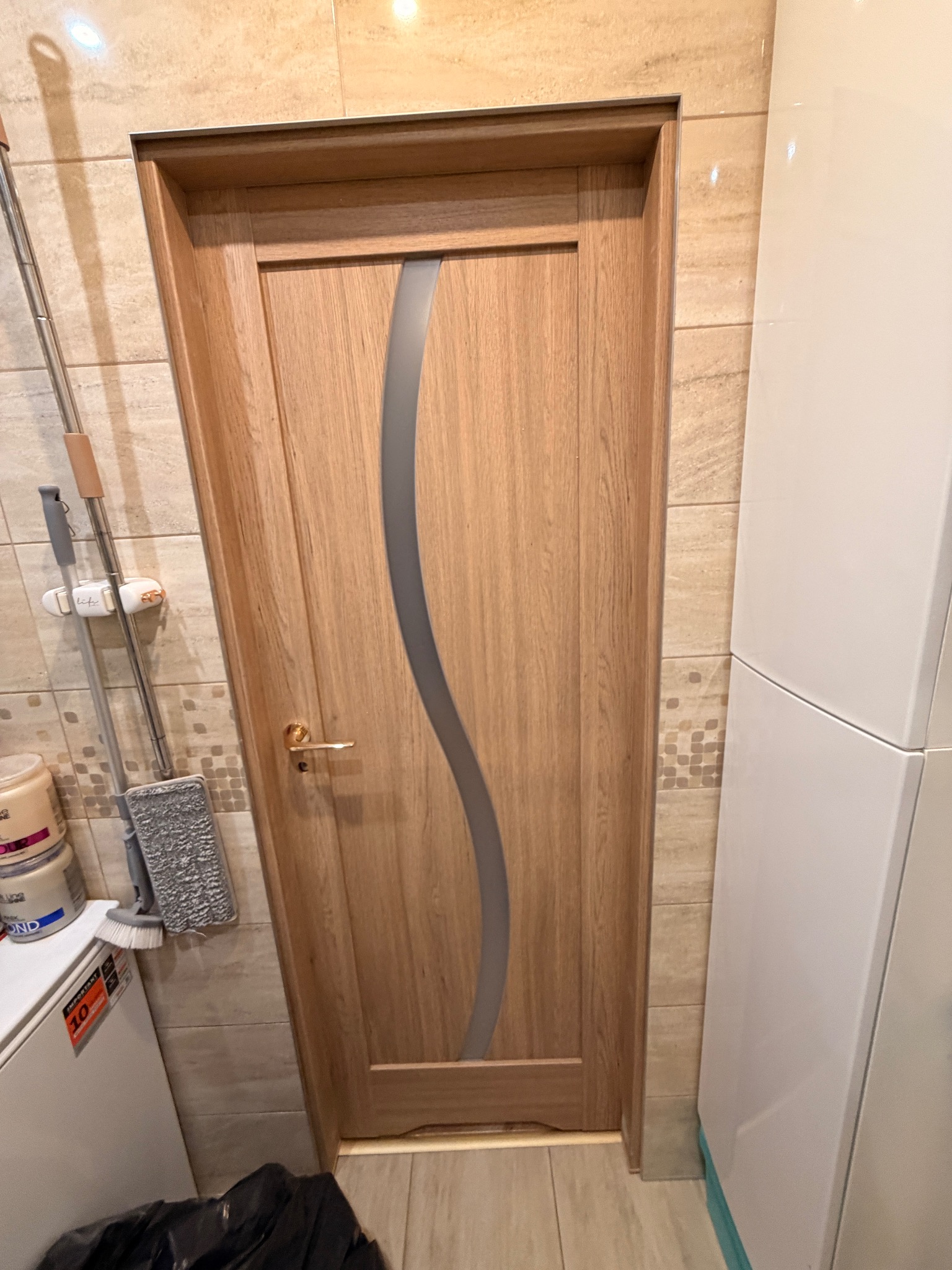 Drewniane drzwi wewnętrzne z falistym, szarym przeszkleniem, widoczne w łazience z beżowymi płytkami. Złota klamka, obok mop i szafka.