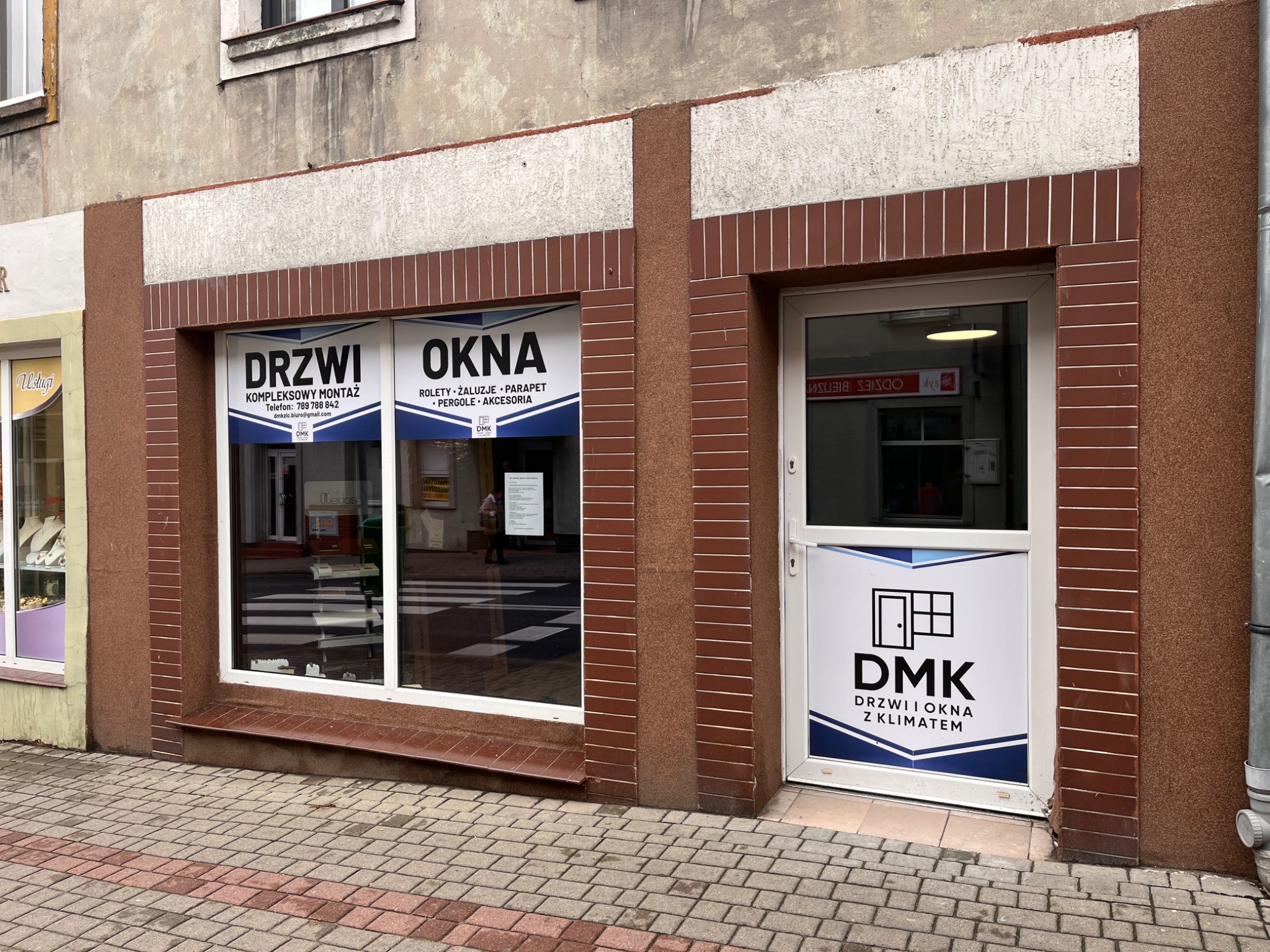 Fasada lokalu usługowego z witryną i wejściem, na których umieszczono reklamy firmy DMK oferującej drzwi i okna. Widoczne szyldy z ofertą i danymi kontaktowymi.