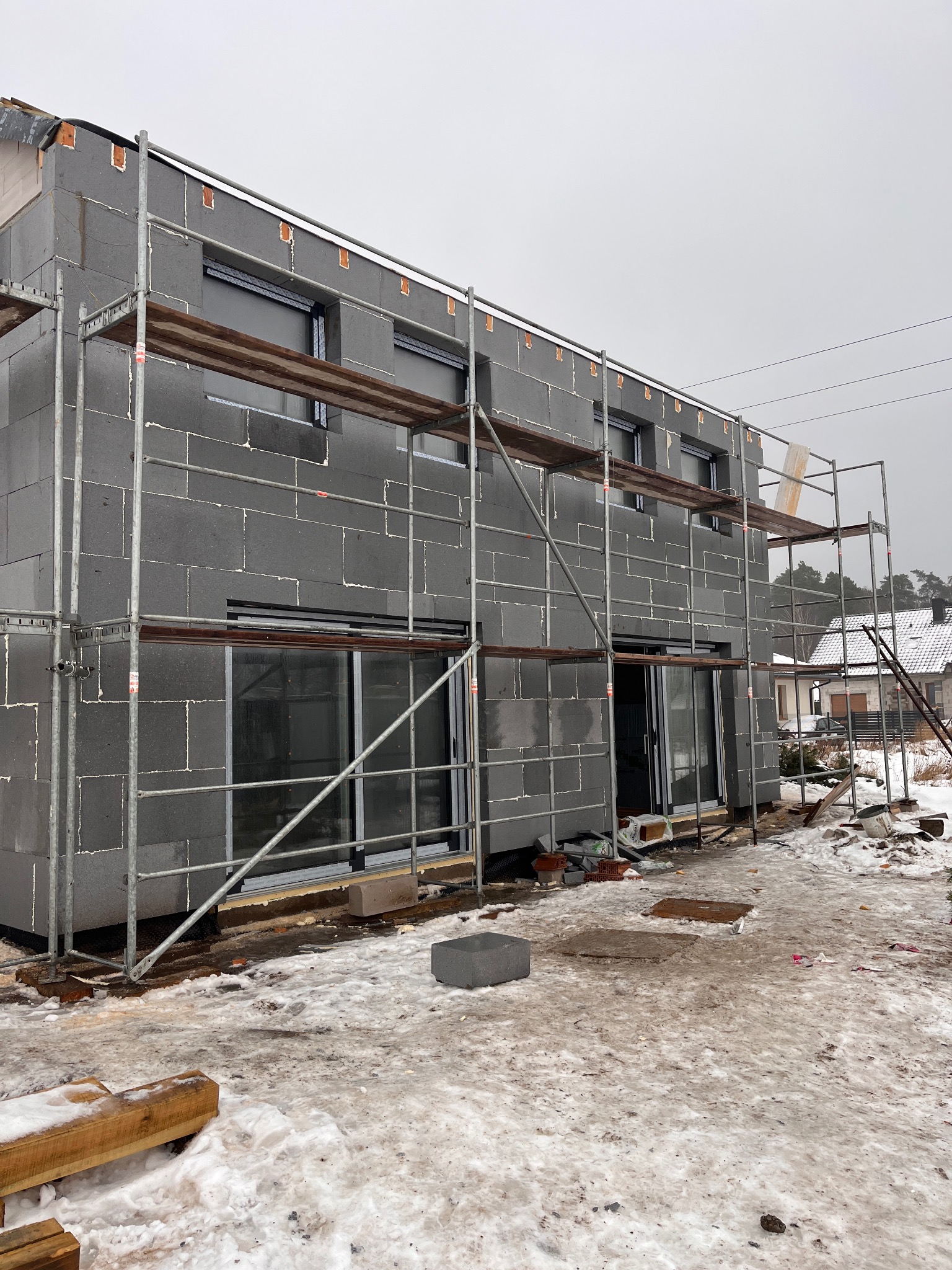 Ocieplanie budynku z szarego styropianu, widoczne rusztowania i zamontowane okna. Prace w toku, zimowa sceneria z resztkami śniegu.