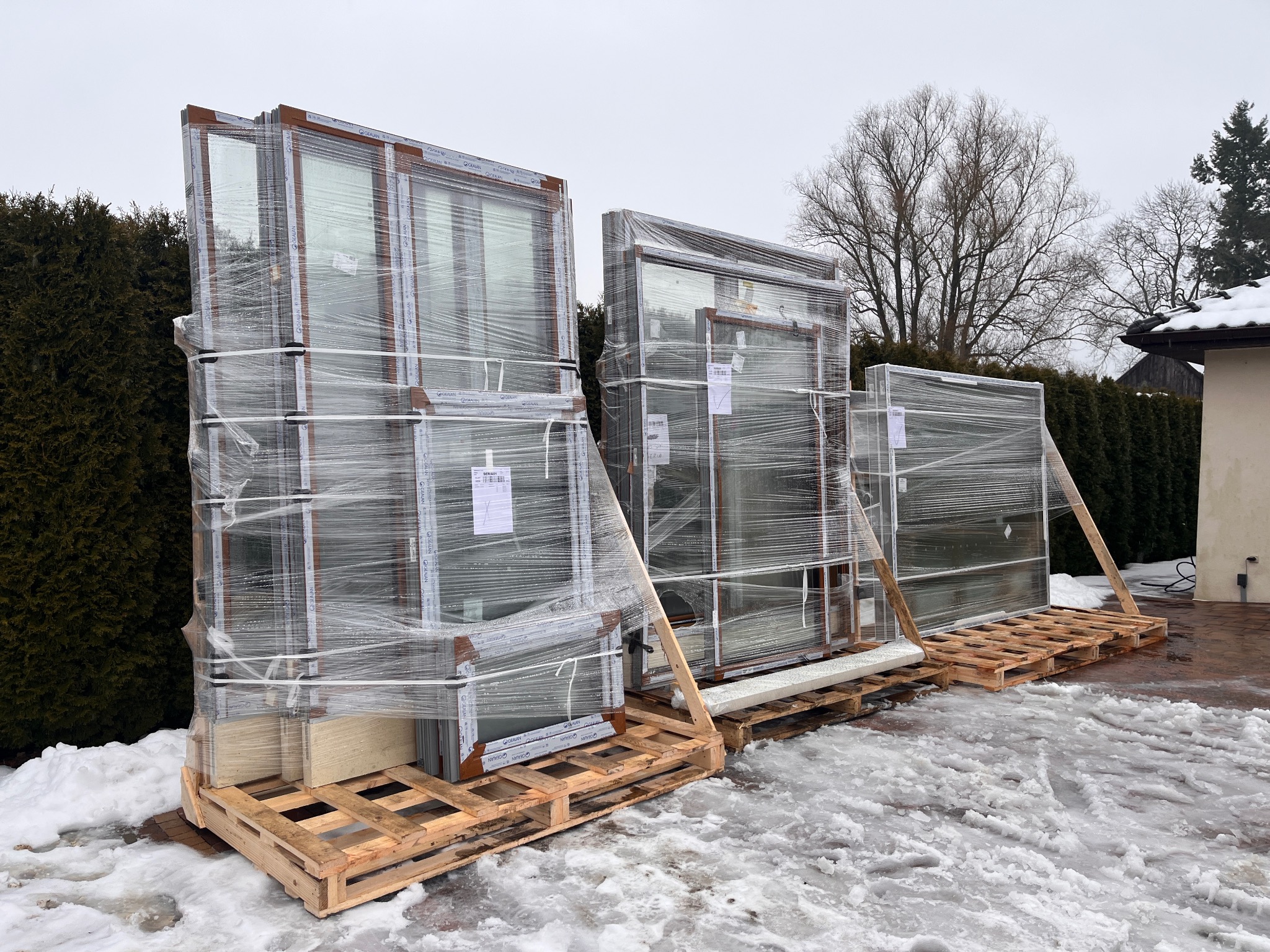 Pakiety okien z brązowymi ramami, zabezpieczone folią na drewnianych paletach, leżą na śniegu przed budynkiem i zielonymi krzewami. Widoczne etykiety na oknach.