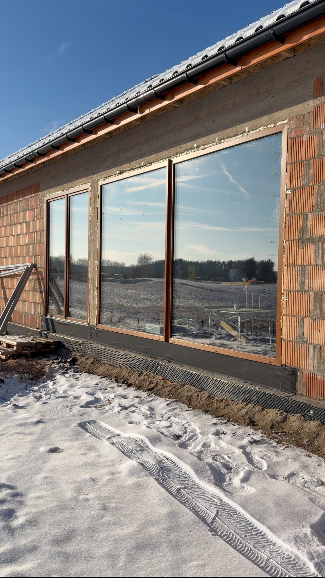 Montaż okien z brązowymi ramami w surowym budynku z cegły. Widoczne zimowe otoczenie i odbicie krajobrazu w szybach. Częściowo ośnieżony teren.