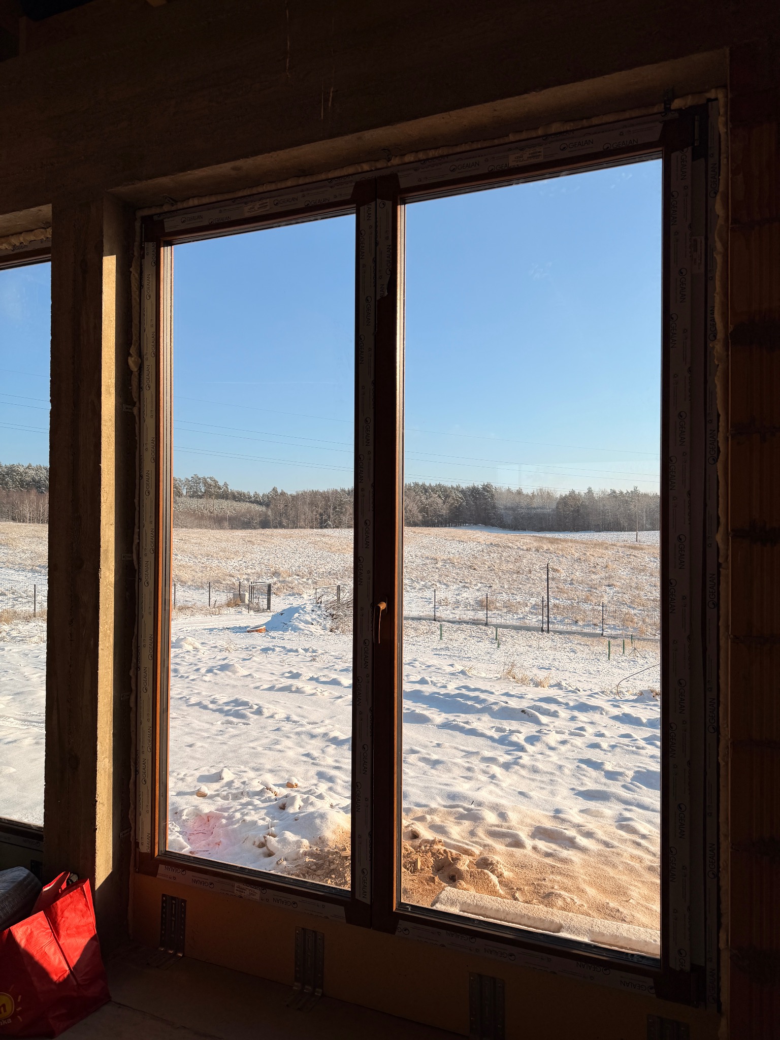 Nowe okno z brązowymi ramami w surowym wnętrzu, widok na zimowy krajobraz z lasem w tle. Elementy montażowe widoczne u dołu ramy. Marka Gealan na profilach.