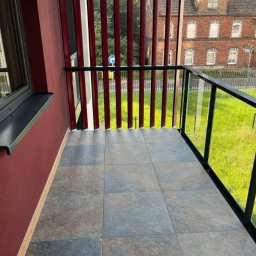 Kamil Patalong - Wykończony balkon z szarymi płytkami podłogowymi, czarną balustradą z elementami szkła i czerwonymi pionowymi panelami. W tle budynek mieszkalny i zieleń.