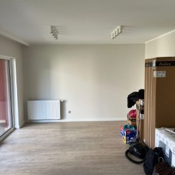 Kamil Patalong - Jasne wnętrze pokoju z panelami podłogowymi, grzejnikiem i kartonami sprzętu AGD. Widoczne lampy sufitowe i duże okno balkonowe. Ściany w odcieniach beżu.