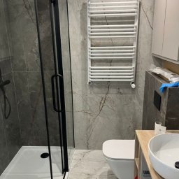 Kamil Patalong - Wykończona łazienka z kabiną prysznicową z czarnymi profilami, grzejnikiem drabinkowym i umywalką nablatową na szafce z jasnego drewna. Płytki imitujące marmur.