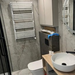 Kamil Patalong - Nowoczesna łazienka z marmurowymi płytkami, wiszącą toaletą, grzejnikiem drabinkowym i szafką z umywalką. Widoczne elementy wykończenia i armatura.