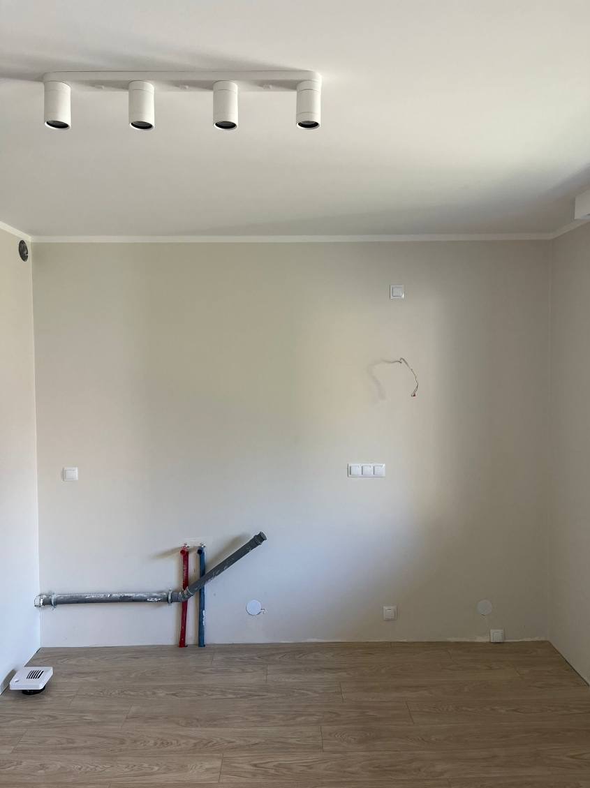 Wnętrze w trakcie prac wykończeniowych: widoczne instalacje wodno-kanalizacyjne przy ścianie, surowy stan ścian i podłoga z paneli. Oświetlenie sufitowe.