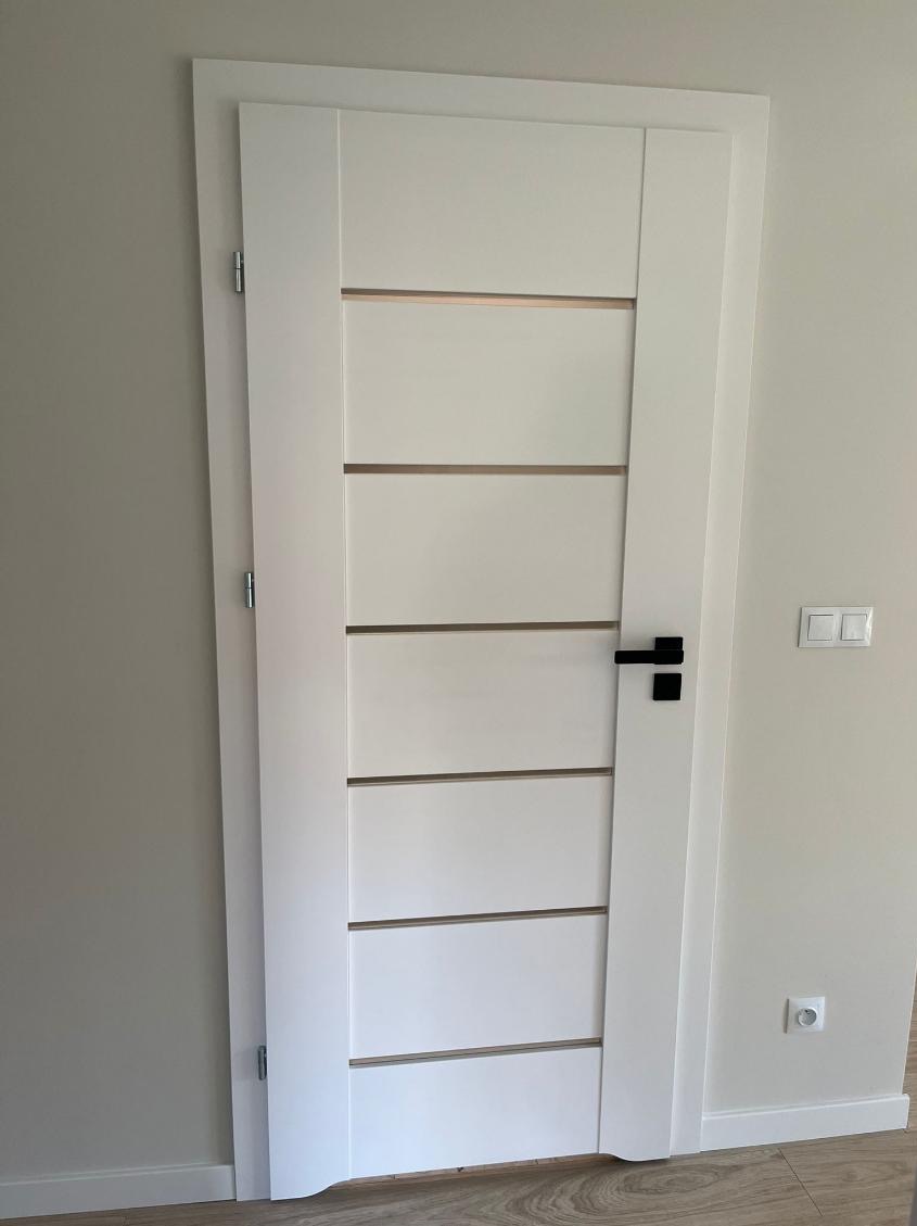 Białe, nowoczesne drzwi wewnętrzne z poziomymi przeszkleniami i czarną klamką, zamontowane w jasnym pomieszczeniu. Widoczne detale wykończenia i gniazdko elektryczne.