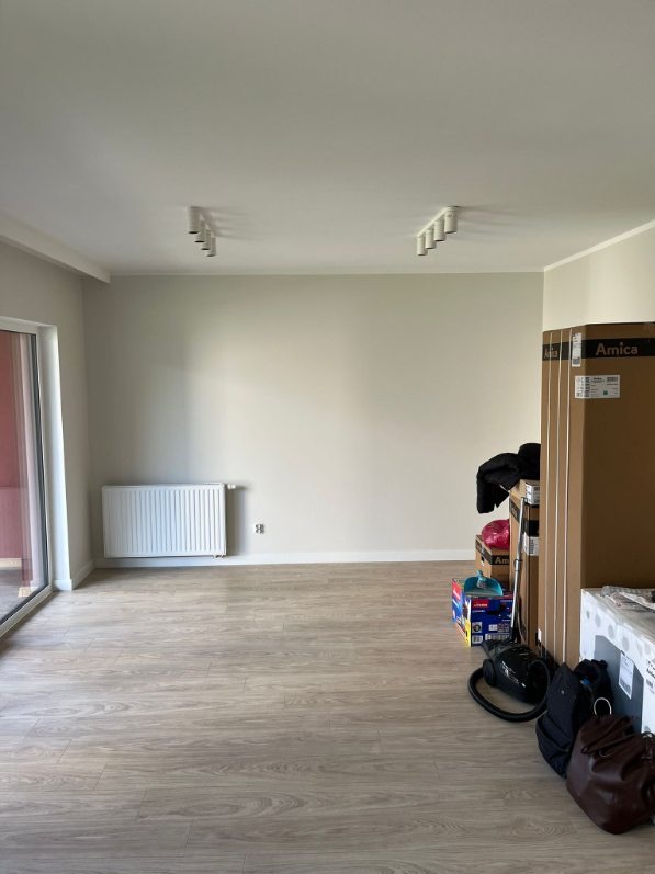 Jasne wnętrze pokoju z panelami podłogowymi, grzejnikiem i kartonami sprzętu AGD. Widoczne lampy sufitowe i duże okno balkonowe. Ściany w odcieniach beżu.