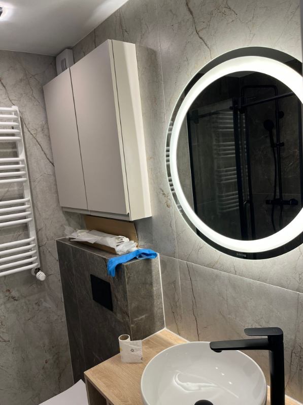 Nowoczesna łazienka z okrągłym lustrem LED, białą szafką i umywalką na drewnianym blacie. Widoczne szare płytki imitujące marmur i grzejnik drabinkowy.