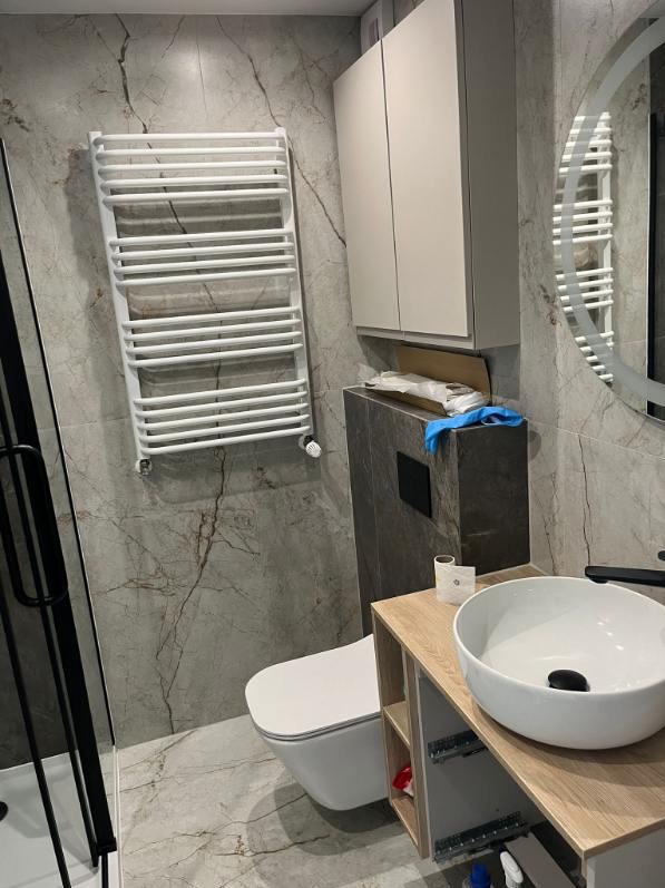 Nowoczesna łazienka z marmurowymi płytkami, wiszącą toaletą, grzejnikiem drabinkowym i szafką z umywalką. Widoczne elementy wykończenia i armatura.