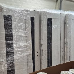 Mb Perfect - Rząd zapakowanych w folię białych drzwi wewnętrznych z czarnymi wstawkami, ustawionych w magazynie. Widoczne otwory na klamki i zawiasy.