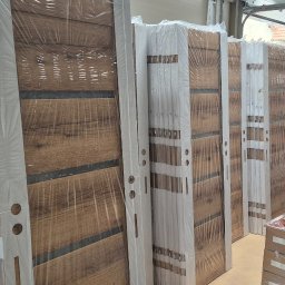 Mb Perfect - Rząd zapakowanych, nowoczesnych drzwi wewnętrznych z drewnianym wzorem, ustawionych w magazynie. Widoczne białe ościeżnice i folia ochronna.