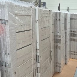 Mb Perfect - Rząd zapakowanych w folię, białych drzwi z poziomymi, ciemnymi paskami, ustawionych w magazynie. Widoczne kartony z napisem 'Góra'.