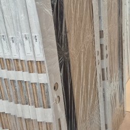 Mb Perfect - Rząd zapakowanych, gotowych do montażu drzwi wewnętrznych w magazynie, widoczne otwory na klamki i zamki, niektóre w folii ochronnej, inne z widocznym wzorem drewna.