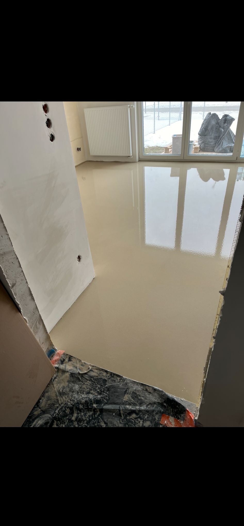 Wylana, gładka posadzka samopoziomująca w jasnym odcieniu beżu, odbijająca światło z okna. Widoczne przygotowane otwory na drzwi i grzejnik.