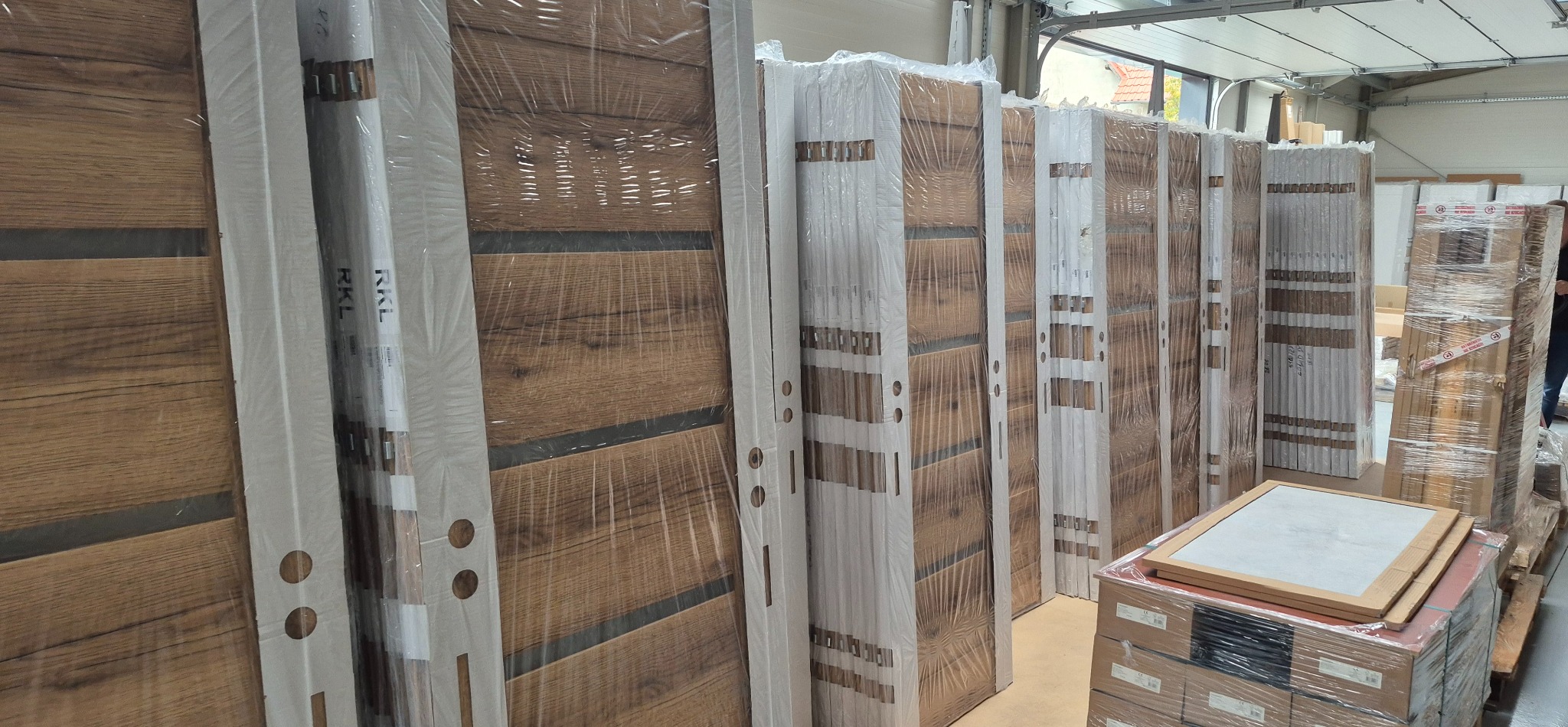 Rząd zapakowanych, nowoczesnych drzwi wewnętrznych z drewnianym wzorem, ustawionych w magazynie. Widoczne białe ościeżnice i folia ochronna.