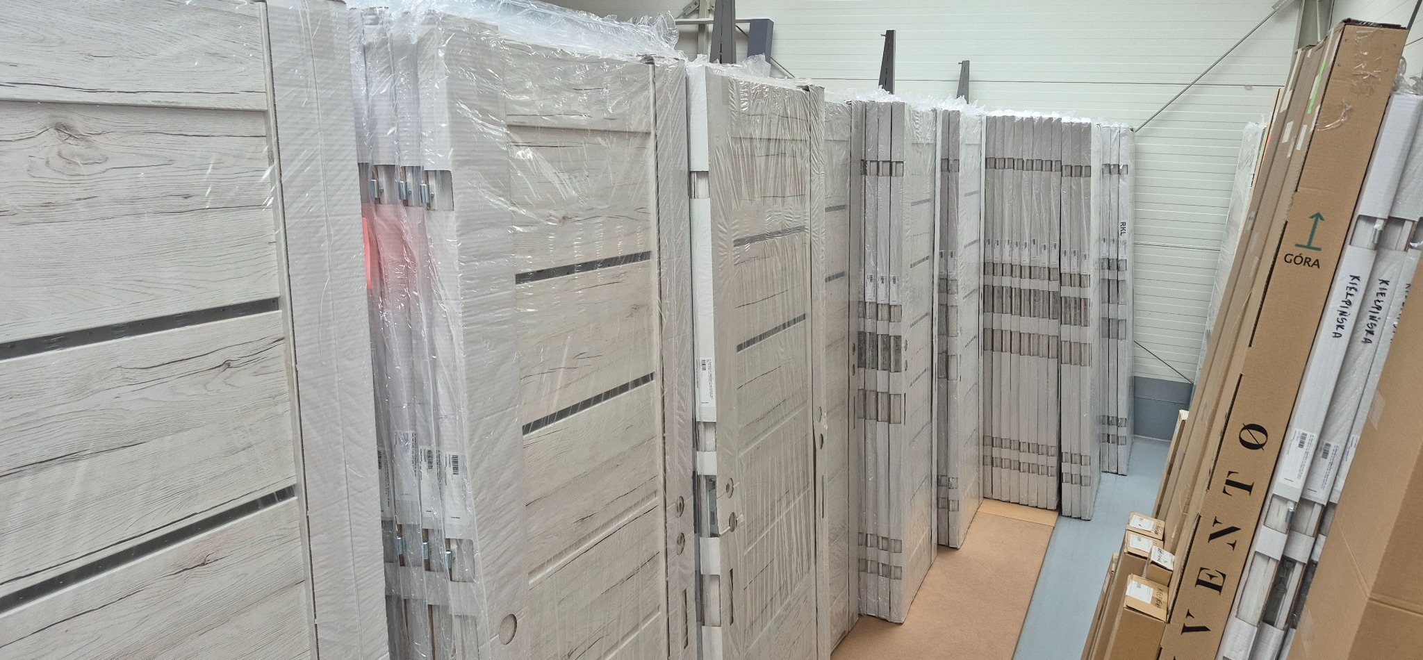 Rząd zapakowanych w folię, białych drzwi z poziomymi, ciemnymi paskami, ustawionych w magazynie. Widoczne kartony z napisem 'Góra'.