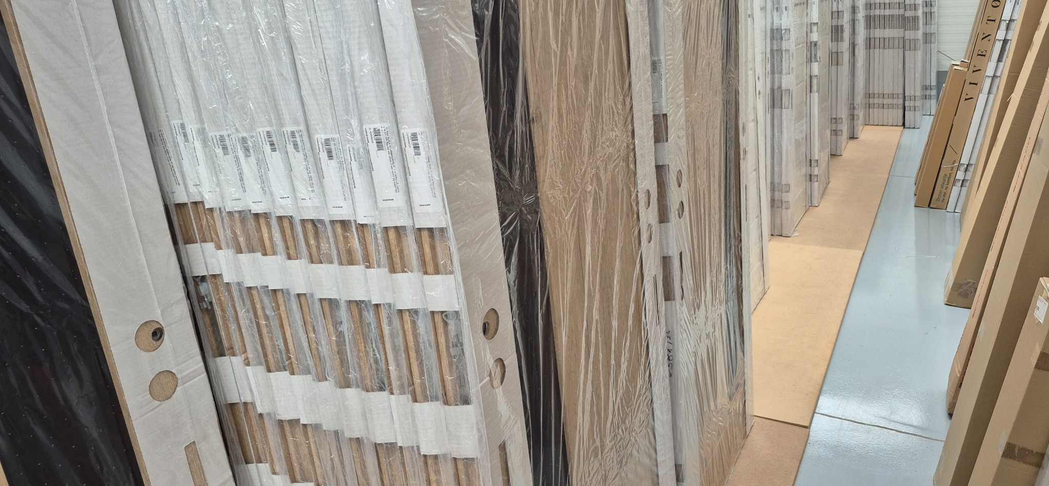 Rząd zapakowanych, gotowych do montażu drzwi wewnętrznych w magazynie, widoczne otwory na klamki i zamki, niektóre w folii ochronnej, inne z widocznym wzorem drewna.