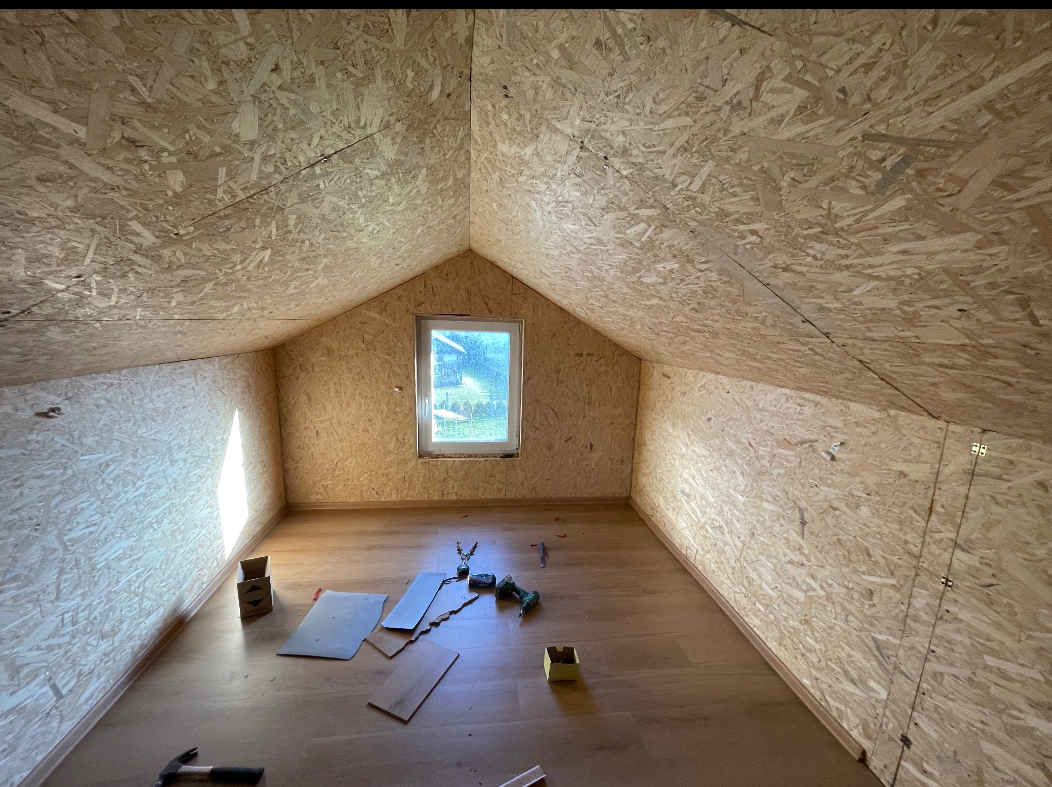 Poddasze z oknem w trakcie wykańczania: ściany i sufit wyłożone płytami OSB, na podłodze panele, widoczne narzędzia i akcesoria montażowe. Ujęcie z dołu, szeroki kąt.