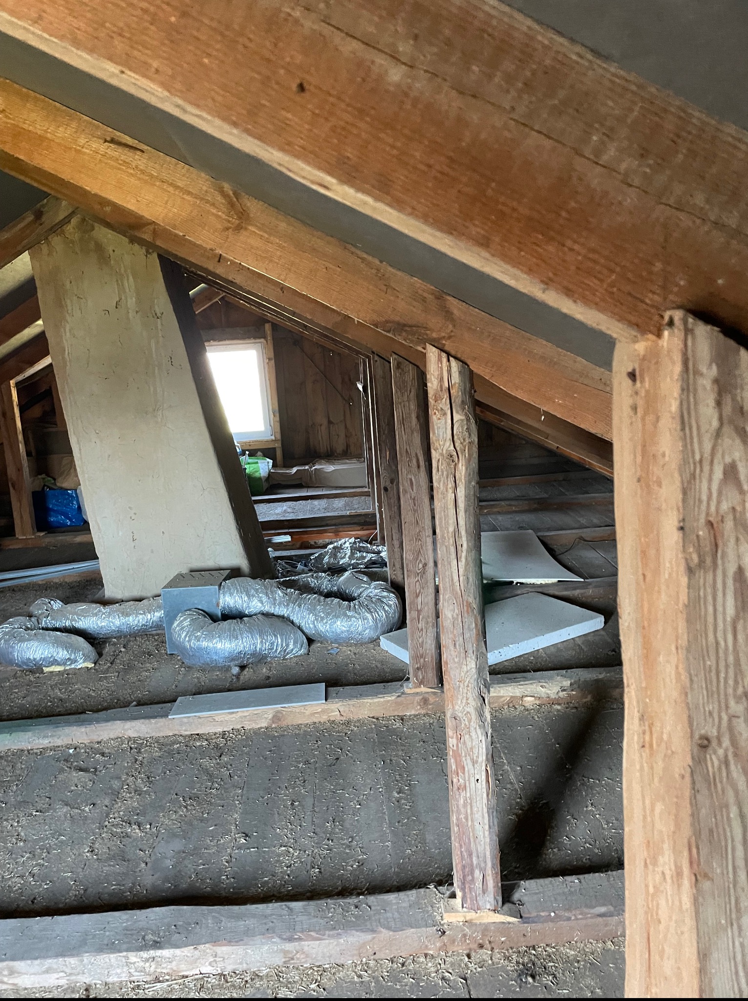 Poddasze w trakcie prac remontowych: widoczne drewniane belki konstrukcyjne, fragment komina, okno oraz elementy instalacji wentylacyjnej z metalowych rur.