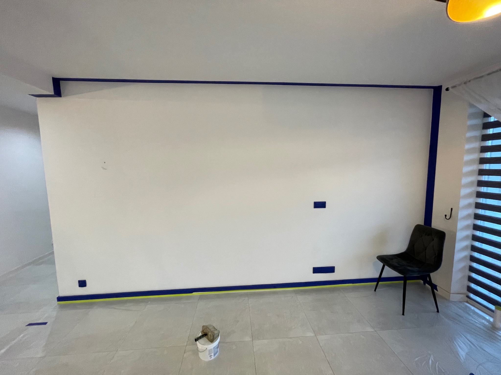 Biała ściana zabezpieczona niebieską taśmą malarską przed malowaniem. Na podłodze wiadro z farbą i krzesło. Przygotowanie do malowania w jasnym wnętrzu.