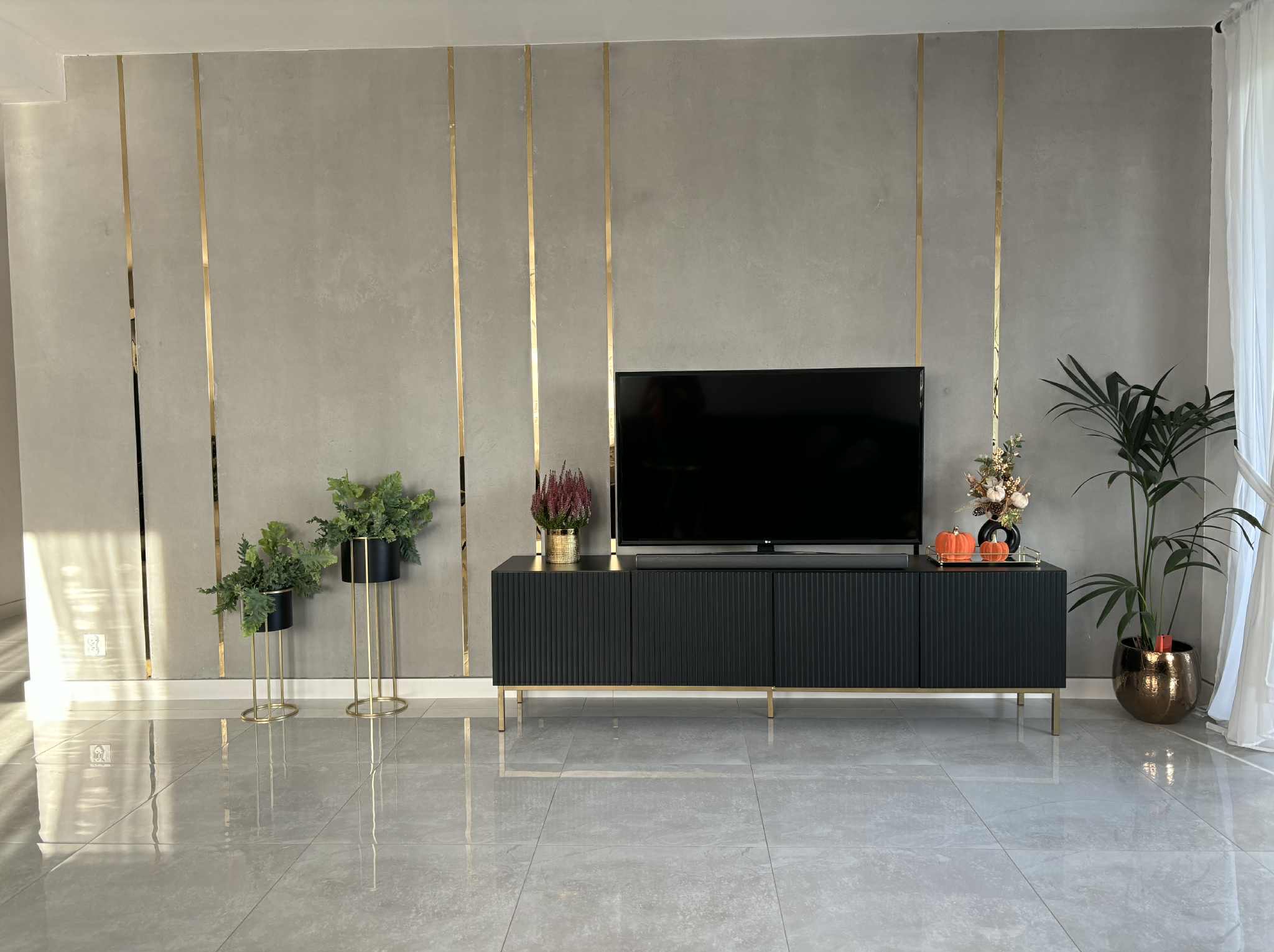 Stylowe wykończenie wnętrza: szara ściana z złotymi listwami, czarna szafka RTV na złotych nóżkach, rośliny doniczkowe. Nowoczesny design, gładkie płytki podłogowe.