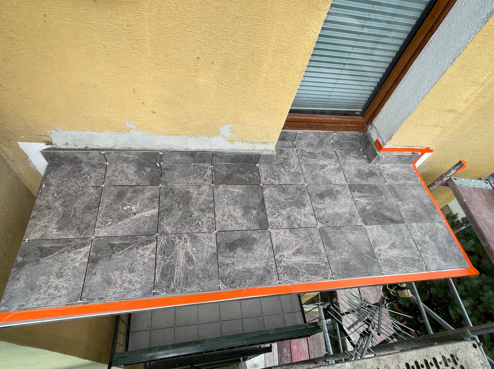 Ułożone szare płytki gresowe na balkonie, widok z góry. Pomarańczowe profile wykończeniowe. Fragment rusztowania w tle. Okno połaciowe w górnej części kadru.