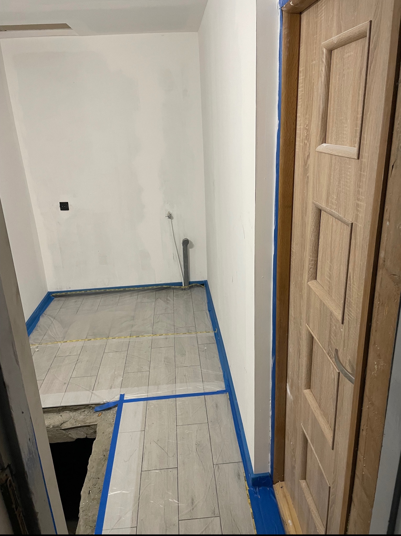 Ułożone płytki podłogowe w łazience, zabezpieczone niebieską taśmą malarską. Widoczna drewniana ościeżnica drzwiowa. Ściany przygotowane do malowania.