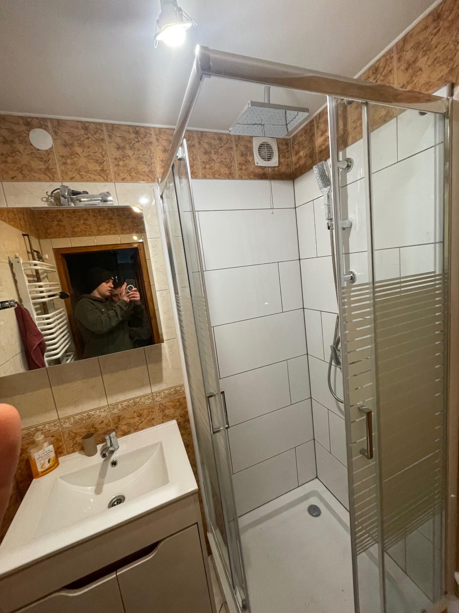 Nowoczesna kabina prysznicowa z białymi płytkami w łazience z beżowymi akcentami. Widoczna umywalka z szafką i lustro z odbiciem osoby robiącej zdjęcie.