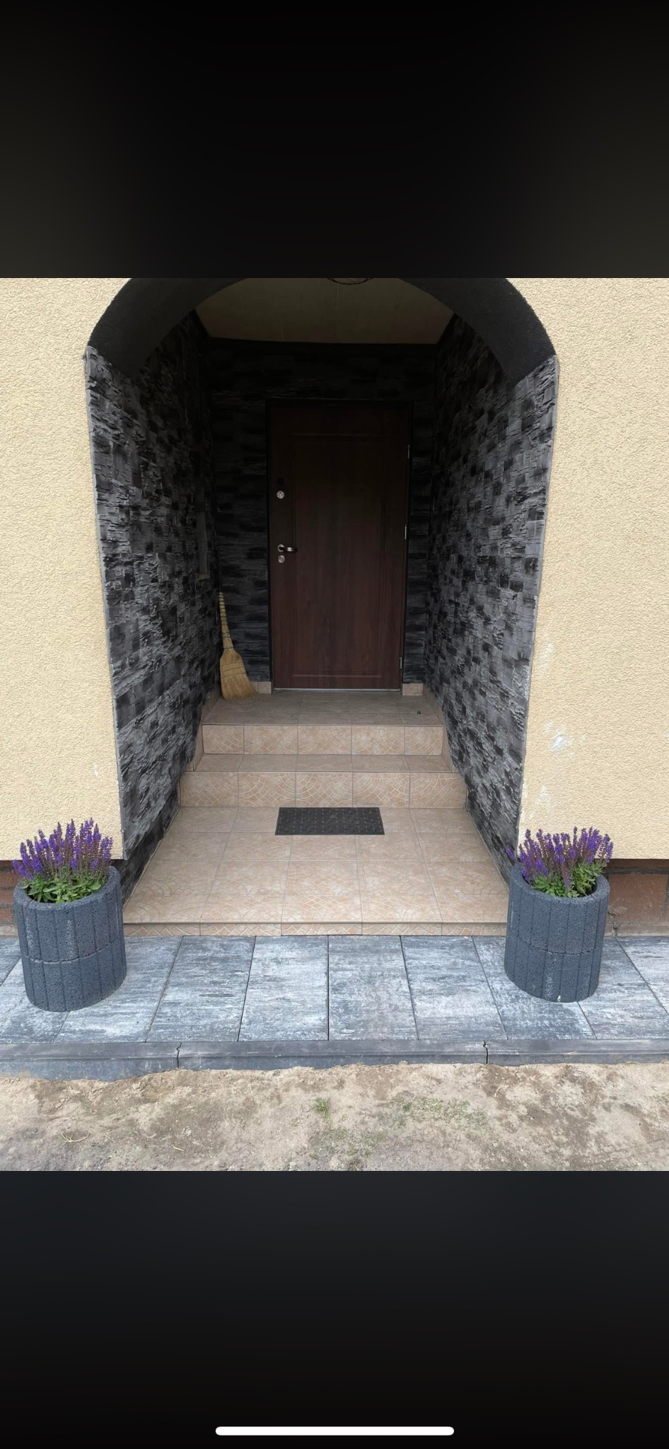 Wejście do domu z kamienną okładziną ścian, schodami z płytek i donicami z fioletowymi kwiatami. Zamiataczka oparta o ścianę.