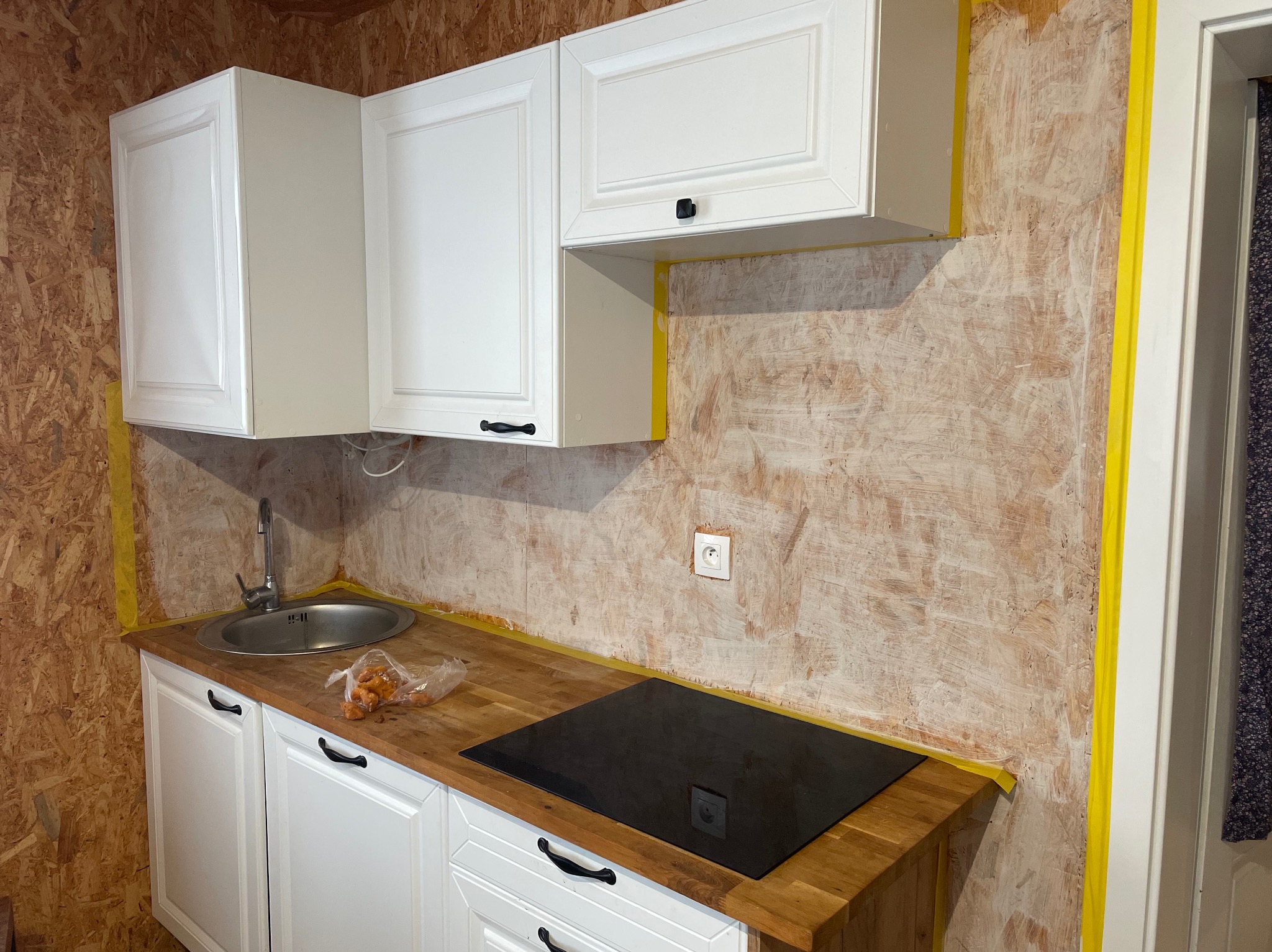 Białe szafki kuchenne z czarnymi uchwytami na drewnianym blacie, zlew i płyta indukcyjna. Ściany przygotowane do malowania, zabezpieczone żółtą taśmą malarską.