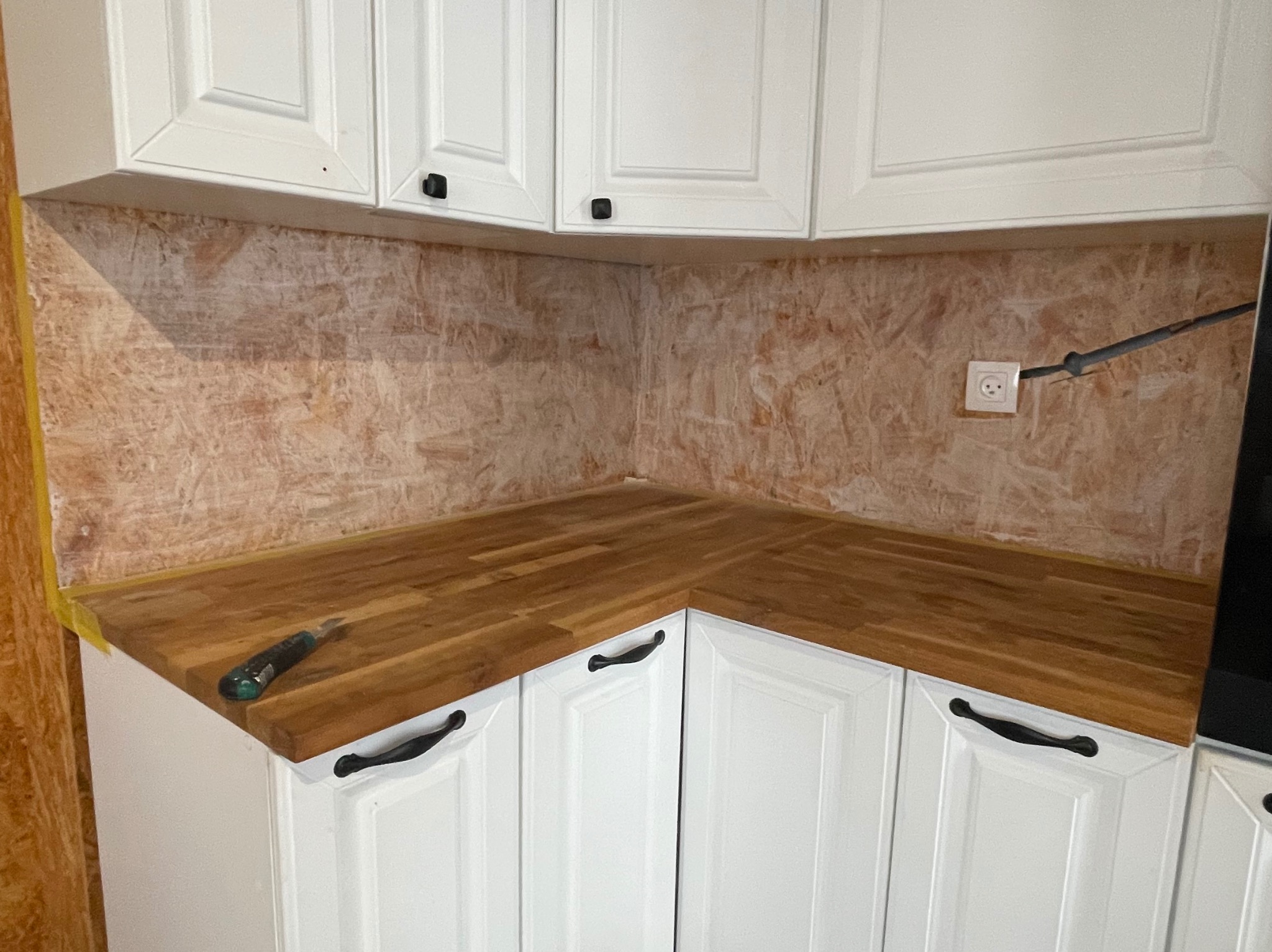 Montaż blatu drewnianego w kuchni z białymi szafkami i przygotowaną ścianą pod płytki. Widoczny nóż na blacie i gniazdko elektryczne.