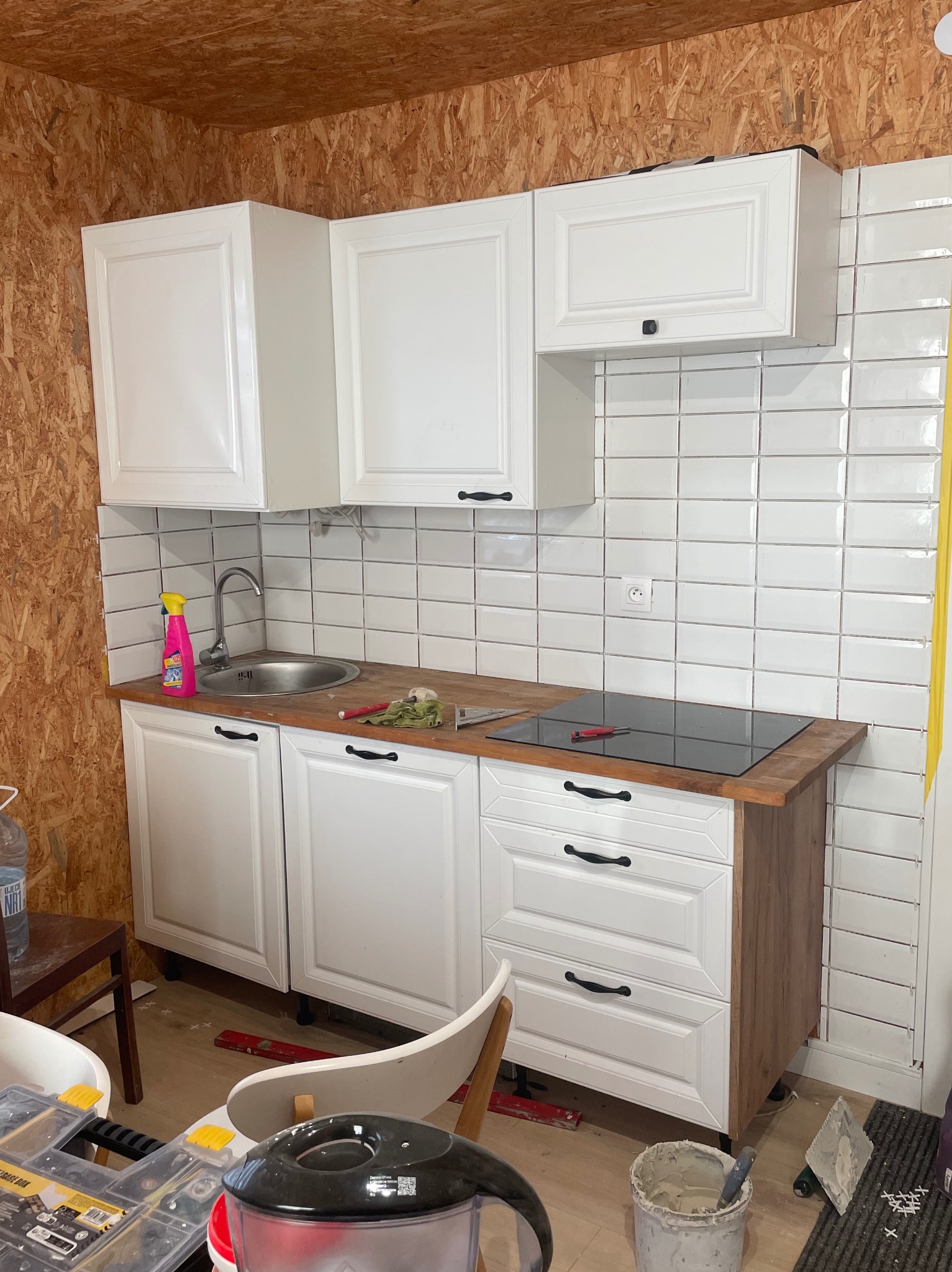 Białe szafki kuchenne z drewnianym blatem i płytkami na ścianie. Widoczne narzędzia i materiały budowlane, co sugeruje remont kuchni w trakcie realizacji.