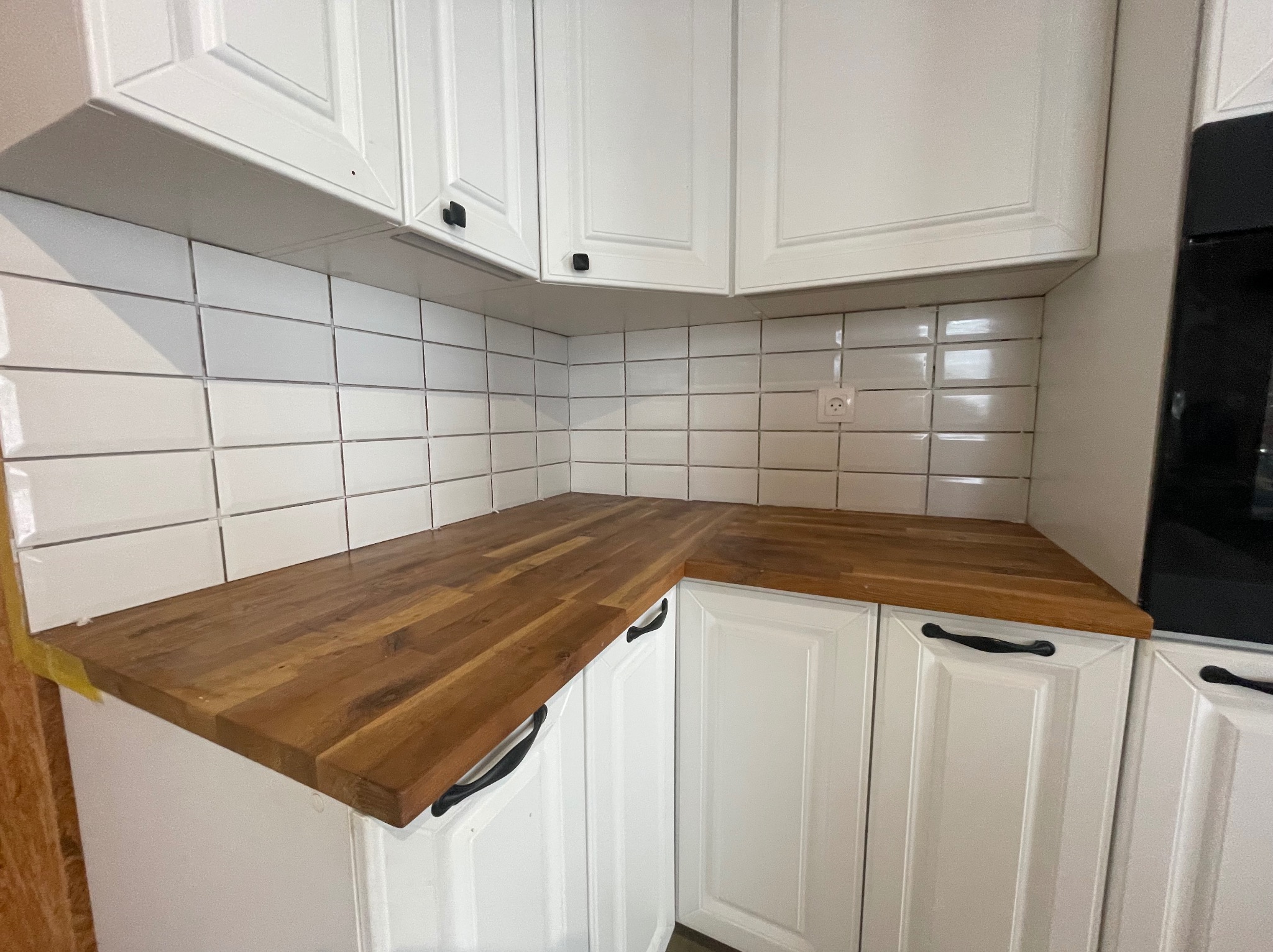 Kuchenny blat z drewna, białe szafki z czarnymi uchwytami i białe płytki na ścianie. Widoczny fragment piekarnika. Ujęcie z bliska, prezentujące detale wykończenia.