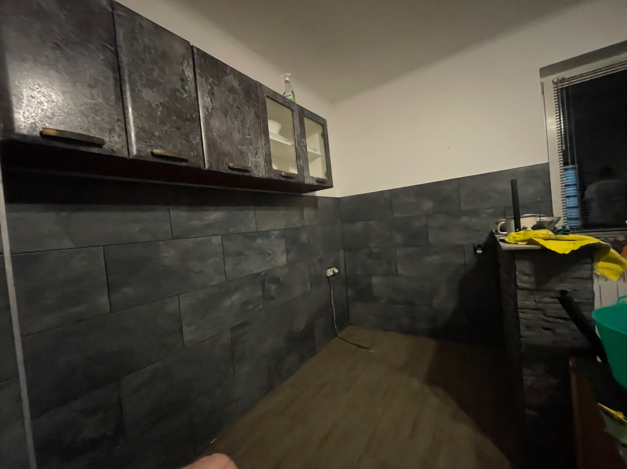 Ciemna kuchnia z szafkami o teksturze betonu i szarymi płytkami imitującymi kamień na ścianie. Widoczny fragment okna i podłogi.