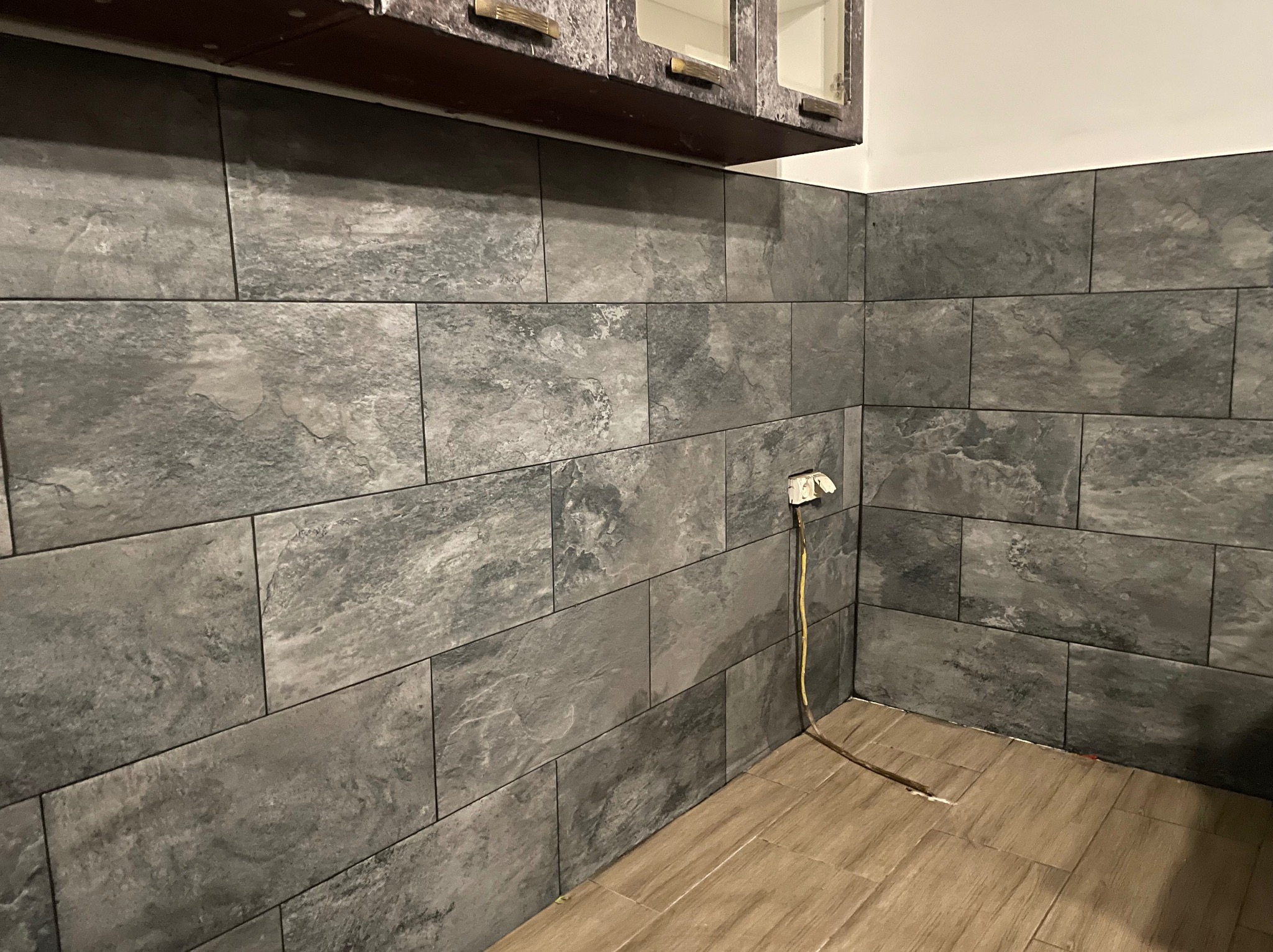 Wykończona ściana w łazience z szarych, matowych płytek imitujących kamień, z widocznym gniazdkiem elektrycznym i fragmentem drewnopodobnej podłogi.