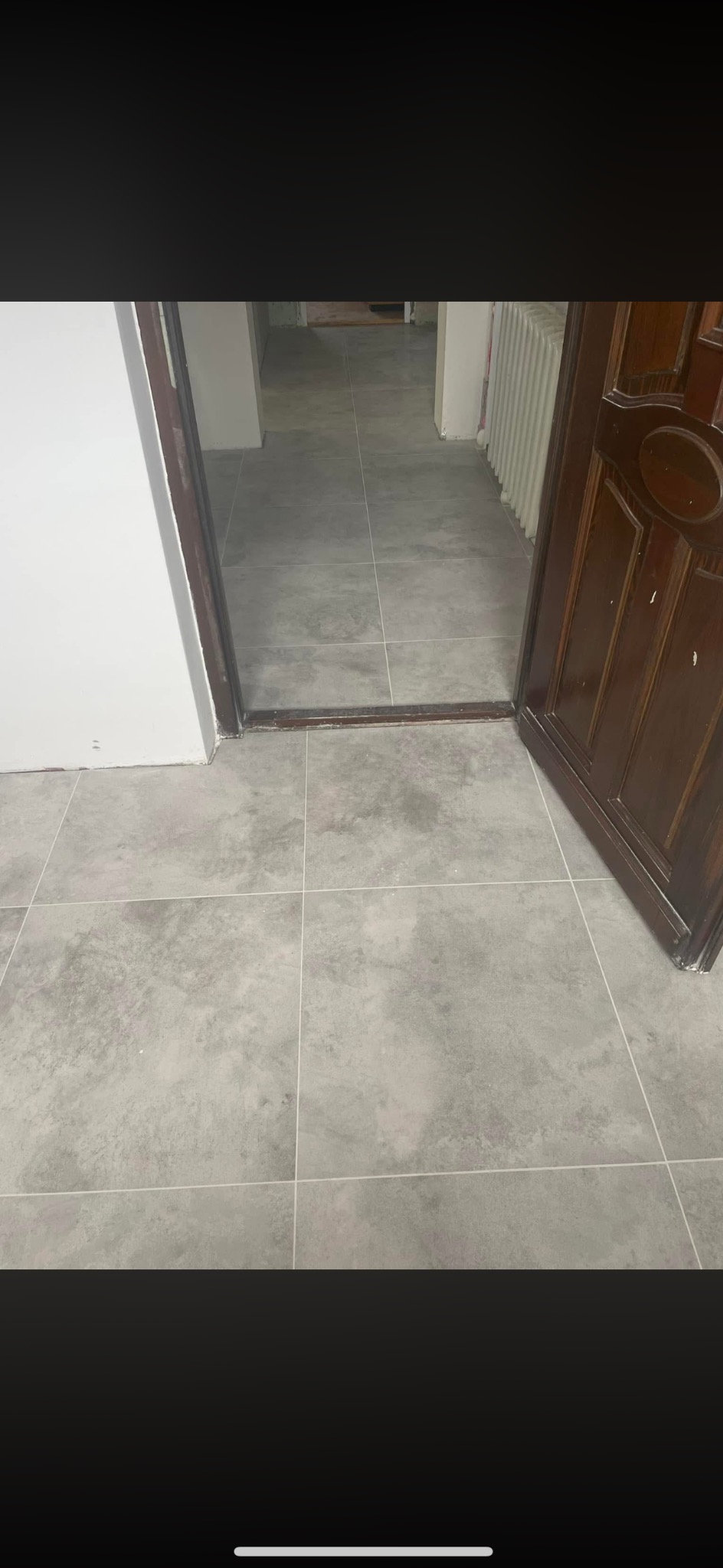 Szare, kwadratowe płytki podłogowe ułożone w korytarzu, widoczne przez otwarte, drewniane drzwi. W tle grzejnik i fragment ściany. Minimalistyczny design.