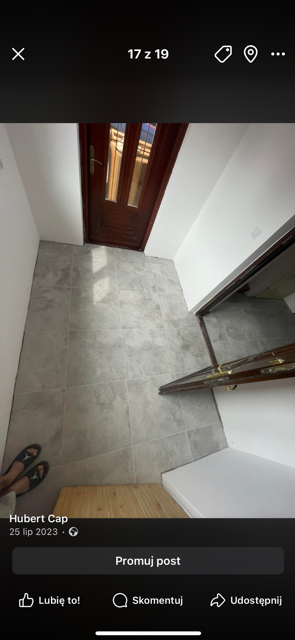 Wykończony korytarz z szarymi płytkami podłogowymi i drewnianymi drzwiami wejściowymi. Widoczne stopy w klapkach. Ujęcie z góry, podkreślające wzór płytek.