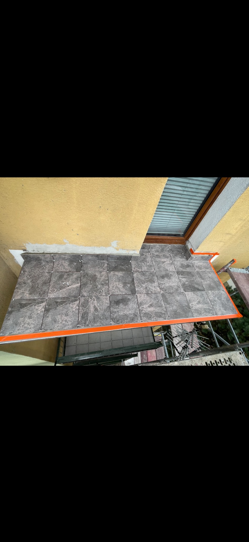 Wykończony balkon z szarymi płytkami imitującymi marmur, zabezpieczony pomarańczową listwą. Widok z góry, fragment rusztowania w tle. Żółta elewacja budynku.