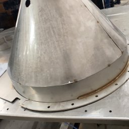 WLD Technology Stanisław Strózik - Stożkowy element metalowy z otworem i śladami spawania, umieszczony na metalowym stole. Widoczne spawy i otwory montażowe. W tle osoby i elementy warsztatu.