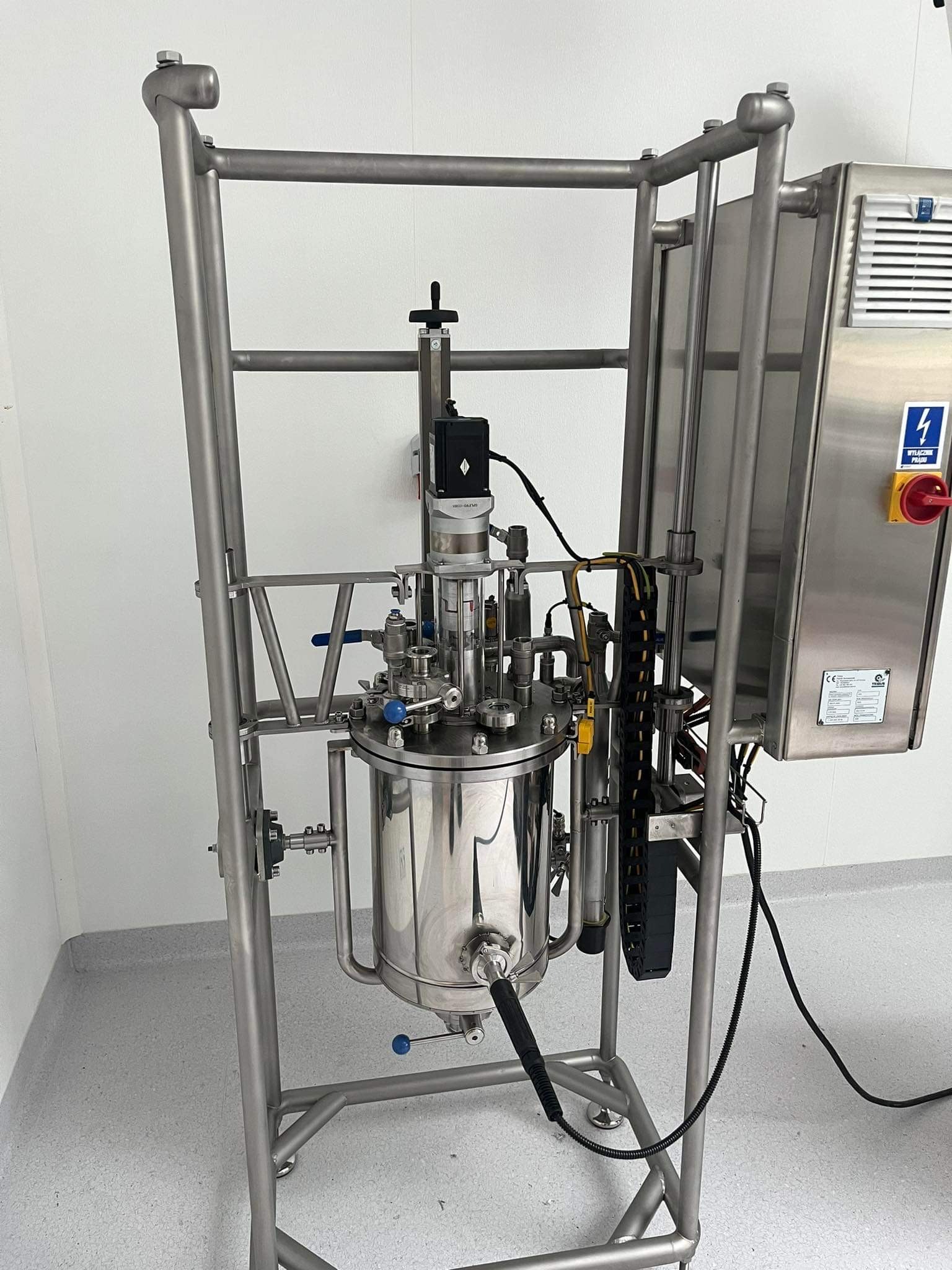 Metalowa konstrukcja wsporcza z reaktorem chemicznym ze stali nierdzewnej, widoczna aparatura kontrolna i panel sterowania. Całość w sterylnym pomieszczeniu.