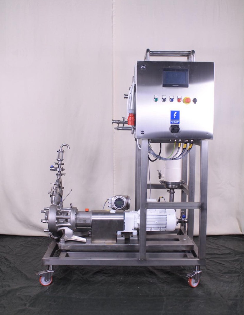 Stalowa konstrukcja z pompą, silnikiem i panelem sterowania na kółkach. Widoczne złącza, przewody i elementy instalacji. Tło stanowi jasna tkanina.