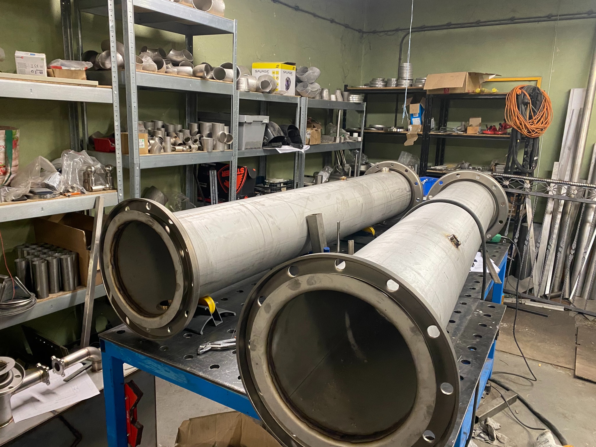 Dwa duże, spawane rurociągi ze stali nierdzewnej z kołnierzami na niebieskim stole warsztatowym w warsztacie, z półkami pełnymi złączek i narzędzi w tle.