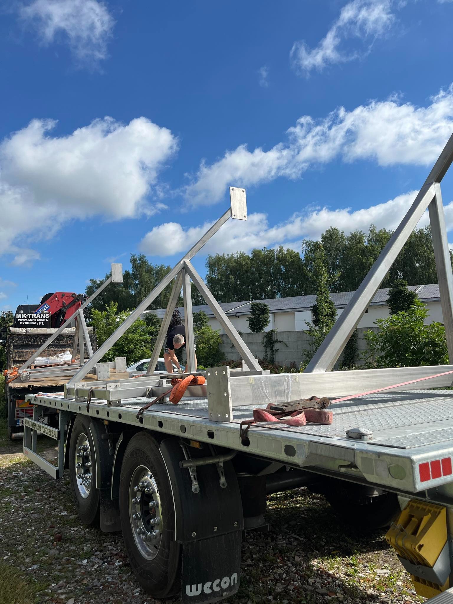 Transport konstrukcji stalowej na platformie ciężarówki, zabezpieczonej pasami. Widoczne elementy spawane i mocowania. Błękitne niebo w tle.