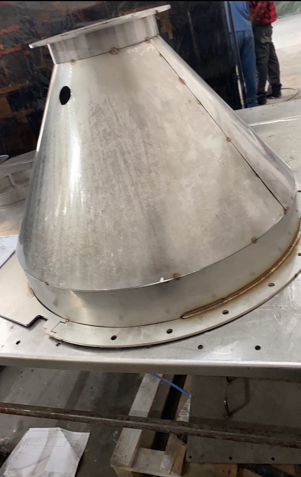 Stożkowy element metalowy z otworem i śladami spawania, umieszczony na metalowym stole. Widoczne spawy i otwory montażowe. W tle osoby i elementy warsztatu.