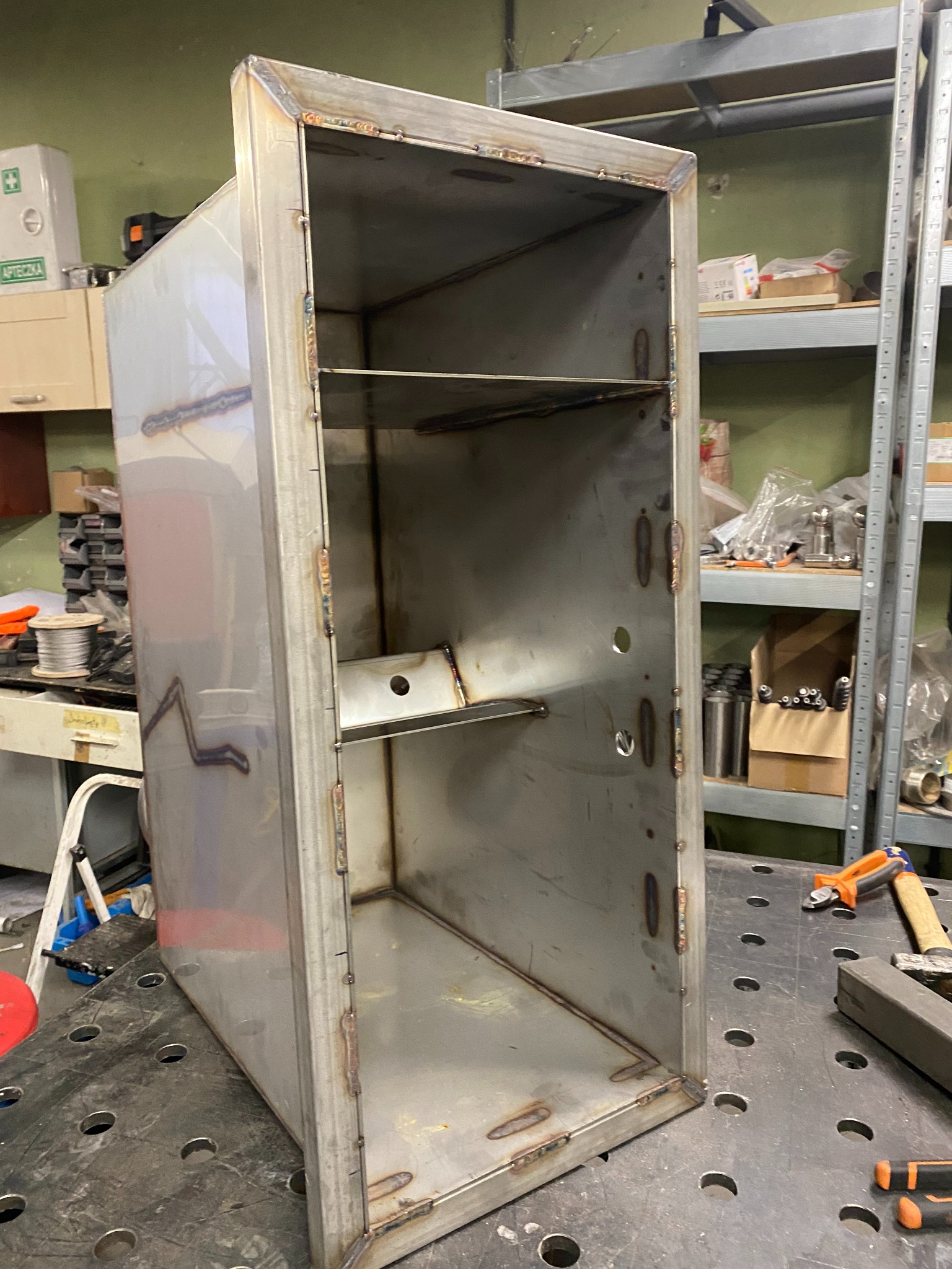 Spawana metalowa szafka z półkami, widoczne spawy na krawędziach, surowy metal, warsztatowe otoczenie, narzędzia na stole.